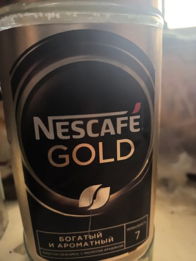 The image shows a close-up of a glass jar of Nescafé Gold coffee. The jar has a black label with gold and white lettering. The prominent text on the label reads "NESCAFÉ GOLD." Below this, there is text in Russian which appears to describe the coffee as "rich and aromatic" and mentions "golden roast with arabica." A numerical rating of "7" for intensity is also visible. The jar is positioned in what appears to be an indoor setting, with a blurred background. There is no visible indication of the city of Orsha, Belarus, or any other specific location.