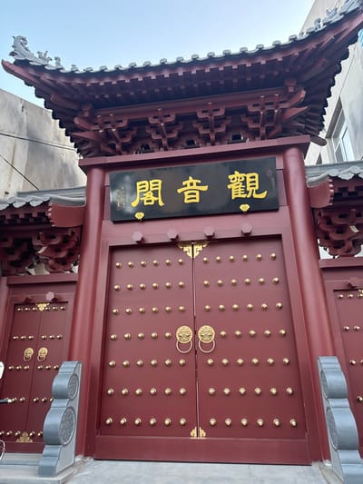 A close-up shot focuses on an ornate, reddish-brown Chinese gate. The gate is constructed with numerous golden studs and decorative door knockers. A black signboard with golden, vertical Chinese characters hangs to the left of the gate, indicating the gate's name. The architectural details showcase traditional Chinese craftsmanship with layered wooden eaves and intricate carvings. The scene is likely located in Jinzhou, China, given the traditional Chinese architectural style. The angle suggests the photographer is standing below, looking upwards towards the gate. The environment appears to be urban, based on the glimpses of adjacent buildings.