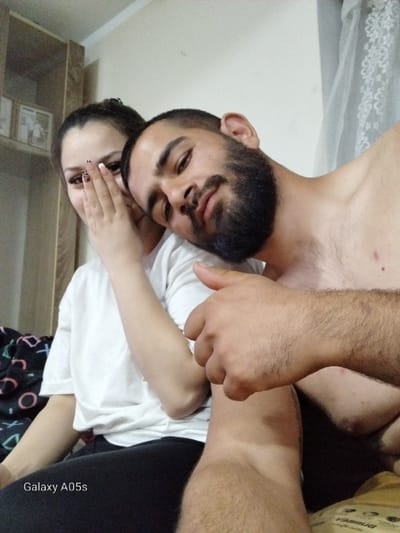 This image captures two individuals, a female and a male, in an indoor setting within the city of Oreshene, Bulgaria. The female, positioned on the left, wears a white t-shirt and dark leggings, with dark hair styled back. Her right hand is raised, covering her mouth and lower face, exposing nails with a black and white leopard print pattern. The male, positioned to her right, is shirtless, with short dark hair and a full beard. His head is tilted and rests on the female's shoulder or upper chest. His right arm is visible, with his hand making a thumbs-up gesture. Both individuals appear to be looking towards the camera. The background on the left includes a light-colored shelving unit displaying framed items. To the right, a light-colored wall with a sheer white curtain is visible. A dark patterned blanket or sheet is partially visible beneath the female on the left. The bottom left foreground displays a "Galaxy A05s" watermark. The individuals are seated or reclining on what appears to be a bed or soft surface.