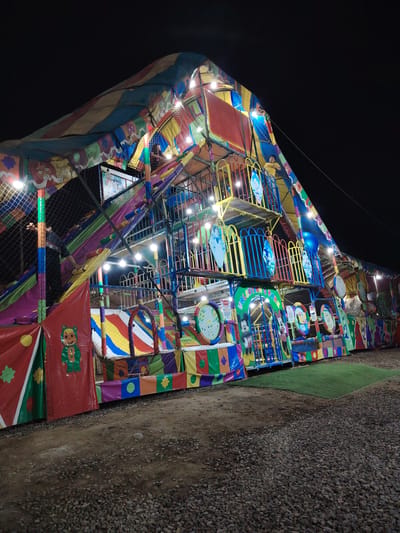 The image shows a brightly colored, multi-level indoor playground structure, illuminated by multiple lights. It is likely located outdoors at night in Trujillo, Peru. The structure includes slides, climbing areas, and various decorative panels featuring cartoon characters. Several people are visible within the structure. The ground around the playground is covered in gravel, with a small patch of artificial turf visible to the right of the structure.