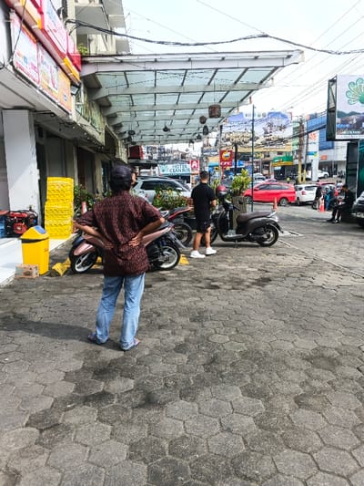 This image depicts a busy urban street scene in Bien Hoa, Vietnam, observed from a paved sidewalk under an overcast sky.
In the foreground, an individual is seen from behind, facing away from the viewer. They wear a dark baseball cap, a patterned brown and black short-sleeved shirt, light blue jeans, and blue flip-flops, with their hands on their hips. The sidewalk is paved with hexagonal grey concrete blocks, showing some damp or discolored areas. To the left of this individual, against a building, are a yellow trash receptacle with a blue lid, a stack of yellow plastic crates, and a red portable generator-like object. A light pink scooter is parked to the left of the foreground person.
Slightly further into the midground, a second individual, partially obscured from the rear, wears a black t-shirt, black shorts, and white sneakers, standing near a dark grey scooter. An overhead glass canopy extends from the adjacent buildings, supported by metal framework, featuring multiple decorative dark-toned hanging lamps.
Beyond the paved area, a street with vehicular traffic is visible, including a prominent red hatchback, a white SUV, and numerous motorbikes. Buildings lining the opposite side of the street display various commercial signs and billboards, including one clearly stating "HOTEL CIANJUR" and another featuring natural landscape imagery. Utility wires are suspended against the light grey, overcast sky. On the far right, several individuals are visible, some seated near parked motorbikes.
