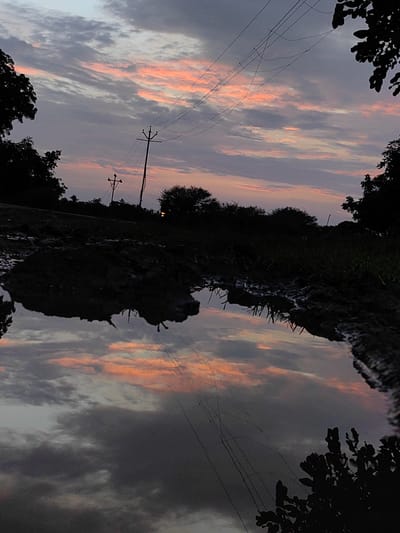 The image captures a muddy puddle reflecting a dusky sky filled with streaks of pink and gray clouds. Silhouetted against the sky are trees, power lines, and a utility pole. The foreground shows the muddy texture of the ground and the dark reflection of the landscape in the water. The scene appears to be in a rural or semi-rural setting, possibly on the outskirts of Dattapur, India, given the presence of overhead power lines and the vegetation. There are no people or animals visible in the frame.