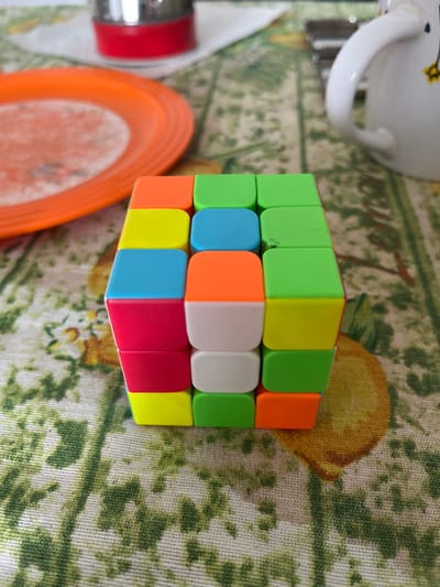 A close-up, eye-level, medium shot shows a colorful Rubik's Cube situated on a patterned tablecloth. The cube is the primary subject, with its sides displaying a variety of colors including orange, yellow, blue, pink, white, and green. The tablecloth features a repeating pattern of green foliage and yellow fruit, possibly lemons. In the blurred background, an orange plate is visible on the left, and a white mug with a dark pattern is on the right. A red and silver cylindrical object is also partially visible in the upper left background. The scene appears to be indoors, on a table. The image does not contain enough information to determine its specific location in Senago, Italy.