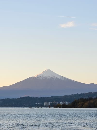 The image captures a landscape scene in Villarrica, Chile. A tall mountain, potentially Volcán Villarrica, dominates the view, its peak partially covered in snow. The mountain's lower slopes appear to be densely forested. In the foreground, a body of water, likely Lake Villarrica, reflects the light. Along the lake shore, there are buildings visible, perhaps residences or hotels. A jet contrail is also seen in the sky.