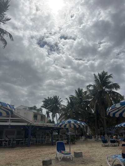 The image shows a beach scene, likely in Le Gosier, France, given the context. The sky is overcast with grey and white clouds dominating the upper left portion of the frame, while a few palm trees and several blue-and-white striped beach umbrellas are visible along the right side. A building with white and light pink walls is also present near the beach. Beach chairs and some people are seen near the umbrellas, indicating a relaxed beach atmosphere. The foreground shows sandy ground, likely the beach itself. The scene overall suggests a coastal area in a tourist location, possibly during cloudy weather.