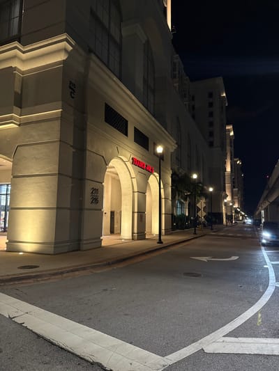 This nighttime image, captured in Coral Gables, United States, depicts an urban street scene featuring a large, multi-story building illuminated by artificial light. The building, likely commercial, showcases a light-colored facade with prominent arches over ground-floor entrances and recessed windows on upper floors. Its architecture suggests a Mediterranean or Spanish Colonial Revival style, characteristic of the area.
A bright red sign prominently displays "TRADER JOES" above one of the arched entrances, identifying a well-known grocery store. Below this sign, numbers "211" and "215" are visible on the building's pillars, likely indicating unit or address numbers. A faint "C" is also discernible on a pillar further to the left. Streetlights positioned along the sidewalk cast warm, directional light onto the building and the adjacent street.
The asphalt street is dark but clearly visible, with distinct white road markings. A large white arrow painted on the road indicates a right turn. The street appears empty of pedestrians and vehicles in the immediate foreground, though blurred lights in the distant background suggest ongoing traffic or city activity. The sky is completely dark, confirming the late hour. The scene is quiet and well-lit, highlighting the building's architectural details and the urban setting.