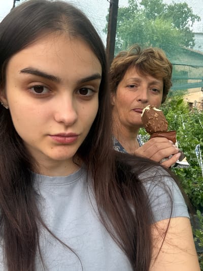 In the foreground of the image, a young woman with long, dark hair is taking a selfie. She is wearing a light blue t-shirt and has a neutral expression. In the background, an older woman with short, brown, curly hair is visible, holding a chocolate-covered ice cream cone and licking it. She is wearing a patterned blue blouse. The setting appears to be outdoors, with greenery and a mesh-like structure in the background, suggesting a garden or greenhouse environment. The image may have been taken in Krupnik, Bulgaria, given the context provided.
