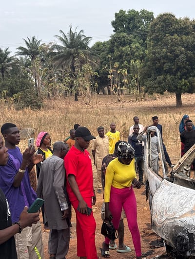A group of people stand in a field next to a burnt-out vehicle. Some are taking pictures with their phones. There are trees in the background. The sky is overcast. The field is dry and grassy. Some of the people are wearing brightly colored clothing, including yellow, red, and pink.