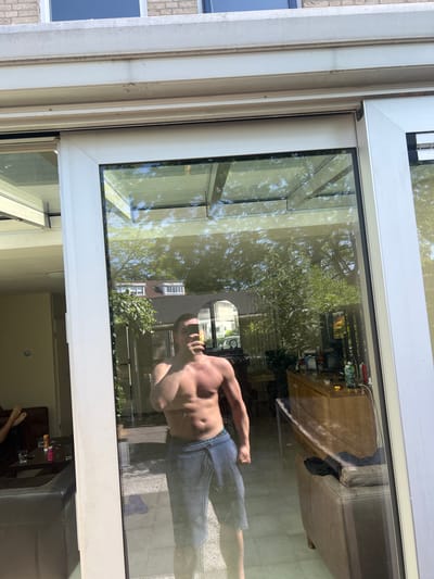 The image shows a shirtless man reflected in a glass door or window. The reflection also shows a tree and a house. Inside, reflected on the window glass, a desk with various objects and a dark couch are visible. The exterior scene reflected includes a house with a distinctive gable roof and white window frames. The presence of typical Dutch architecture, such as gable roofs, the white window frames, and the overall neatness of the houses reflected suggests a location in the Netherlands. The scene appears to be captured during daylight hours. Based on the architectural style, the outdoor foliage, and the reflection of Dutch houses, the image could be potentially from a neighborhood located in Oud-Beijerland. The presence of the residential buildings and greenery suggests the scene takes place in a residential part of the town. The man is standing in an interior location. The image captures a reflection, not a direct view, so details are inferred.
