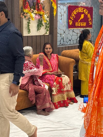 This image captures an indoor scene within Bidhannagar, India, showing several individuals engaged in what appears to be a social or religious gathering. The floor is covered entirely by a plain white sheet.

In the left foreground, a male's torso and legs are visible as he walks from left to right. He wears a dark blue collared shirt with white polka dots, light beige trousers, and a dark wristwatch on his left wrist. His feet are bare.

In the midground, two females are seated on a fluted, camel-colored cushioned sofa. The left-seated woman is elderly, with grey hair and glasses, wearing a pink long-sleeved top and a maroon patterned saree or skirt. She faces away from the camera, seemingly interacting with the woman beside her. Her feet are bare. The right-seated woman is younger, facing slightly towards the camera with a subtle smile. She wears a vibrant pink and red saree or lehenga, the red portion featuring intricate gold embroidery. Multiple gold bangles adorn her wrists, and a red bindi is visible on her forehead. She holds a light-colored mobile phone in her left hand. Her feet are bare.

Behind the seated women, a wall features a white panel decorated with a floral arrangement of pink, purple, and white flowers, interspersed with green leafy garlands. Above these, small red and yellow triangular flags are visible. To the right of this wall panel, a glass partition with a repeating, abstract geometric pattern serves as a background element.

Mounted on the glass partition is a red banner with yellow Hindi text and a golden mace (gada) symbol. The text reads "श्री हनुमान घाटा भक्त मण्डल" (Shri Hanuman Ghata Bhakt Mandal), translating to "Shri Hanuman Ghata Devotee Group/Organization," and below it, "सन्ध्या बाजार" (Sandhya Bazaar), meaning "Evening Market" or "Evening Fair."

To the right of the seated women, a third female stands, mostly with her back to the camera, wearing a yellow salwar kameez-style outfit. Her hair is pulled back in a ponytail. She wears dark blue footwear.

In the far right foreground, a portion of an orange fabric is visible, possibly part of a devotional garment or banner. White Hindi text, specifically "श्री राम" (Shri Ram) repeated, is visible on this fabric.