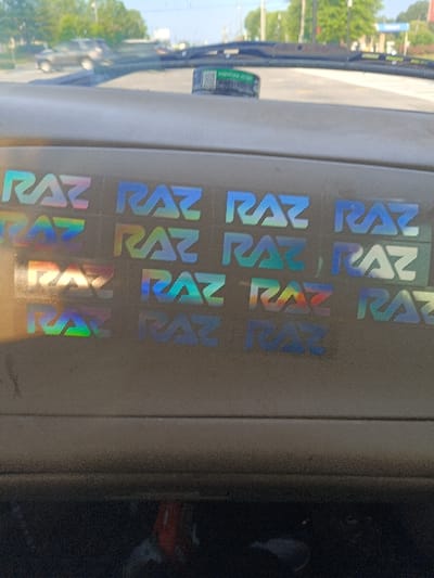 The image shows the inside of a car's windshield. The car is parked at a location in Auburn, Alabama, United States, with coordinates (latitude: 32.623, longitude: -85.448). The car's dashboard is tan. Several iridescent stickers with the word "RAZ" on them, in varying fonts and sizes, are visible on the dashboard. A small, cylindrical object with the word "WARNING" on it is visible on the windshield. The car is parked on a road with a small amount of traffic in the background.