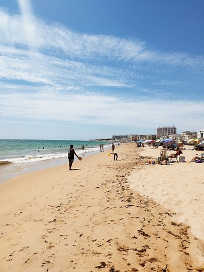 A wide, sandy beach is depicted under a bright blue sky with wispy white clouds. The ocean, a deep turquoise, meets the shore with gentle waves. Several people are visible on the beach and in the water.

In the foreground, a man stands on the wet sand near the water's edge, holding a paddle. Further down the beach, a child is bent over, possibly playing with sand. Scattered along the beach are beach chairs, umbrellas, and bags, indicating leisure activities. In the background, buildings and structures line the coastline, suggesting an urban presence beyond the beach. The sand is imprinted with numerous footprints, showing evidence of recent activity. The overall scene conveys a vibrant and sunny day at a populated beach. This is likely the beach of Rota, Spain, given the urban development visible in the background which is consistent with the city's coastline.