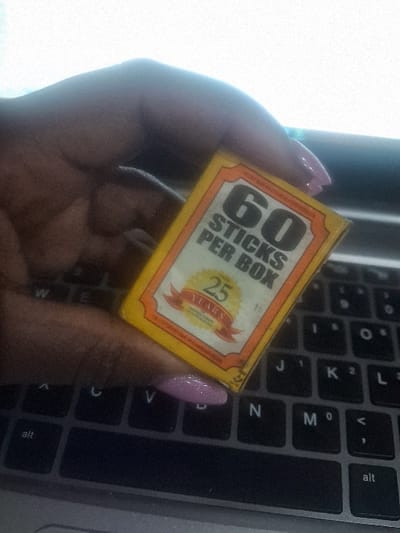 A hand with light purple painted nails holds a small yellow box with orange and white detailing against a black keyboard background. The box reads "60 STICKS PER BOX" in bold lettering. A circular logo on the box declares "25 YEARS" within a red banner and golden ribbon design. The keyboard shows keys like "M", "N", "K", "L", "alt", and "V". A dark cylinder can be seen blurred in the background.