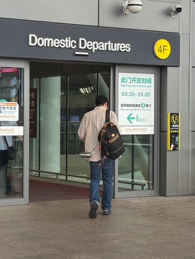 The image shows the entrance to "Domestic Departures" at the 4F gate of Beijing Daxing International Airport, located in Guangyang District, China. A person wearing a plaid shirt, blue jeans, and a backpack is walking away from the camera towards the entrance. To the right of the entrance is a yellow sign with a phone icon and the words "HELP POINT" in English and Chinese, along with some buttons and a finger icon indicating touch activation. Above the entrance, the words "Domestic Departures" are visible in large white letters on a dark gray background. To the right of this, a yellow circle with "4F" is displayed. A sign to the right of the entrance indicates opening hours for the gate from 04:30 to 24:00 and directs people to Gate 3 during closing hours, with an arrow pointing left indicating a distance of 55 meters. A glass door with a red carpet inside is visible.