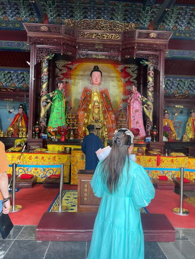 Here is a detailed description of the image:

**Scene:** The image appears to be taken inside a temple or shrine in Lintong District, China. It showcases an ornate altar featuring multiple statues of deities or important figures. The setting exudes an aura of reverence and cultural significance.

**People:** The primary individual is a woman dressed in a traditional light blue outfit and decorative headpiece. She stands with her back to the camera, gazing reverentially at the statues. Her presence indicates she might be praying or observing the shrine.

**Subjects:** The primary subjects are the statues within the altar. They feature detailed clothing and iconography, hinting at their identity as religious or historical figures. Their arrangement and the ornate backdrop create a focal point of the scene.

**Objects:** The altar is adorned with elaborate woodwork, including carved columns, intricate moldings, and decorative panels. Various banners with Chinese characters, lamps, and other religious items are placed strategically around the altar, enhancing its visual appeal and symbolic value.

**Actions:** The woman is engaged in a respectful viewing or prayerful contemplation of the altar and its statues. Other actions, if any, might be happening in the background, but the image focuses on the woman's interaction with the sacred space.

**Interactions:** The key interaction is between the woman and the religious setting. She is clearly engaged with the altar and statues, possibly conveying reverence or seeking spiritual connection.

**Environment:** The temple's interior features a combination of traditional architecture and religious iconography. The lighting is subdued, possibly to enhance the atmosphere of reverence, with bright colors on the banners. The overall environment creates a sacred and culturally rich space.