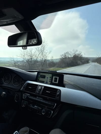 The image depicts the interior of a vehicle, specifically the dashboard and windshield view, on a road outside the city of Piperkovo, Bulgaria.
**Vehicle Interior:** The perspective is from the front passenger seat looking forward. A black steering wheel is partially visible on the left, with a driver's left hand positioned at the 9 o'clock position. The instrument cluster shows analog dials. The central dashboard features an integrated infotainment screen displaying a navigation map interface, indicating a route with an upcoming turn in "500 m" and an instruction like "Take The Drive." The climate control panel below the screen is set to 23.0°C. The dashboard on the passenger side has a textured, light grey trim. A rearview mirror is visible at the top, reflecting an indistinct internal scene. A white cup is present in a cup holder in the lower center. A person's knee or leg is visible in the lower right quadrant, suggesting a passenger.
**Exterior Environment:** Through the windshield, a two-lane asphalt road is visible, stretching into the distance and curving slightly to the right. The road shoulders are unpaved, and the road is bordered by dense, bare deciduous trees and bushes, suggesting a cold season (late autumn, winter, or early spring). Beyond the immediate vegetation, distant rolling fields or hills are visible, with some patches of green. The sky is partially cloudy, with a mix of blue sky and white cumulus clouds.
