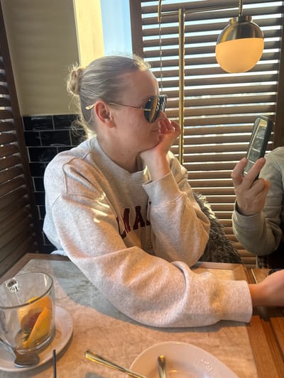 Within an indoor setting in Mysiadło, Poland, two individuals are seated at a light-colored marble-patterned table, illuminated by natural light entering through slatted wooden blinds.
The primary subject is a light-skinned female with blonde hair tied in a bun, facing right and resting her chin on her right hand. She wears gold-framed sunglasses with dark, reflective lenses and a light grey sweatshirt with a dark, possibly maroon, text print on the chest, partially visible as "ALMA". Her left arm rests on the table.
To the right, a second individual's arm and hand are visible, holding a dark-cased smartphone with a visible screen. This person also wears a light grey long-sleeved top. A textured dark grey fabric object, possibly a cushion or garment, is present behind the primary subject.
On the table, a clear glass mug with a handle contains liquid and multiple citrus fruit peels (likely grapefruit or orange), resting on a white saucer with a silver spoon and a dark straw. Adjacent to this, a white plate with visible crumbs holds gold-colored cutlery (a fork and a spoon).
The background features dark wooden slatted blinds, a section of dark, possibly tiled, wall behind the primary subject, and a modern pendant light fixture with a gold-colored top and an opaque white spherical shade.