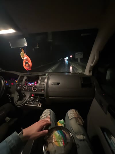 The scene depicts the interior of a vehicle, consistent with a Volkswagen Transporter, during nighttime operation in Gorna Rositsa, Bulgaria. The perspective is from the front passenger seat, looking forward towards the dashboard and the road ahead.
The vehicle's interior is illuminated by the dashboard display lights, which show red and pink accents on the infotainment system and climate control panel. A manual gear shifter is visible on the center console, alongside cupholders and various control buttons, including a hazard light switch. A decorative air freshener, featuring a smiling yellow face above a red, stylized figure, hangs from the rearview mirror. The driver's steering wheel is partially visible on the left.
A passenger occupies the front right seat. The individual's left hand, wearing a light blue long-sleeved garment, rests on a clear plastic container with a colorful, rainbow-striped label on their lap. The passenger wears distressed blue jeans featuring a vertical striped patch on the right thigh and a sepia-toned image patch on the right knee. A small green and white packet is situated near the passenger's right knee.
Outside the vehicle, the asphalt road is wet and reflects distant lights, suggesting rain or recent precipitation. A red circular road sign is visible on the right side of the road, followed by a larger, blank rectangular sign or billboard. The surrounding environment is dark, indicating a rural or suburban thoroughfare.