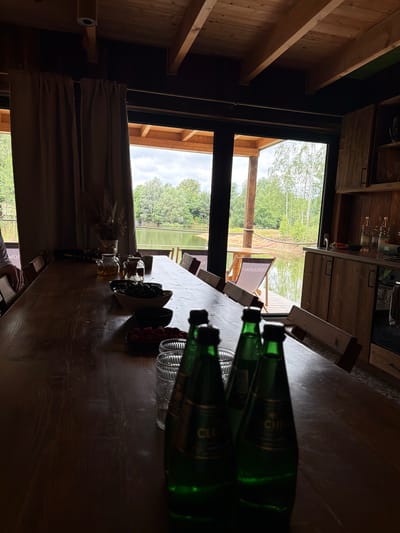 The image depicts an indoor dining area with a large wooden table situated in front of a scenic view of a body of water and trees. The room appears to be rustic, with wooden beams on the ceiling and wooden cabinetry on the right side of the frame. On the table, there are several glass bottles of water, bowls of fruit (cherries and raspberries), and two glass teapots. One person's arm is visible on the left side of the table, resting on a chair. The environment suggests a peaceful setting, possibly a vacation home or a restaurant with a view. The overall lighting is dim, with natural light streaming in from the large windows overlooking the outdoor scenery. The location is specified as Dobrzyń, Poland.
