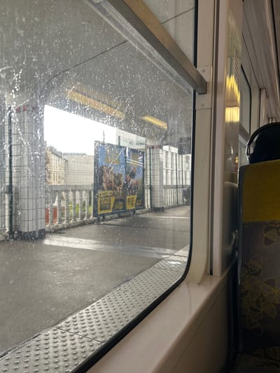 The image depicts a static scene viewed from inside a train or tram at a station platform in Arcueil, France. The perspective is through a window extensively covered with water droplets, indicating rain or a recent cleaning.
On the interior, the right side of the frame shows a portion of a train seat, upholstered in a two-tone fabric: a yellow-green upper section and a dark grey lower section with lighter, abstract, looping patterns. A pair of black over-ear headphones rests on top of this seat. The window frame itself is white or off-white, with a metallic, textured sill featuring raised circular bumps running along its base.
The exterior scene, visible through the wet window, shows a concrete platform that appears damp. Architectural elements include multiple white, grid-patterned structures, possibly support columns or walls, aligned along the platform. A metal mesh fence runs parallel to the track, with several orange traffic cones positioned near its base. In the mid-ground, a large, multi-panel billboard with a predominantly blue background displays animated characters and yellow text banners, likely advertising a film. Beyond the platform structures and billboard, several urban buildings are visible under an overcast or brightly diffused sky. No individuals are present or discernable within the visible exterior.