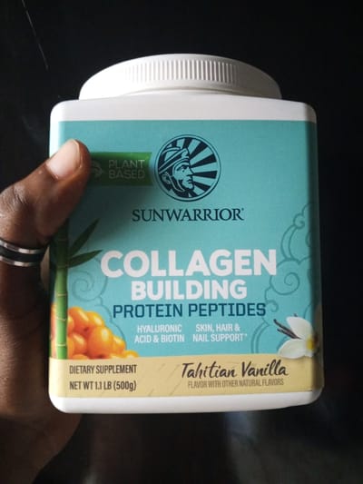 A person's hand, adorned with two silver rings, holds a white plastic container of Sunwarrior Collagen Building Protein Peptides. The container is predominantly teal and yellow. The teal portion features the Sunwarrior logo, a stylized profile of a person wearing a headdress, the words "PLANT BASED," and the product's main title, "COLLAGEN BUILDING PROTEIN PEPTIDES." Below this, smaller text indicates "HYALURONIC ACID & BIOTIN SKIN, HAIR & NAIL SUPPORT*". The yellow band at the bottom of the teal section displays "Tahitian Vanilla" in cursive, with "FLAVOR WITH OTHER NATURAL FLAVORS" beneath it. The lower portion of the container is white and displays "DIETARY SUPPLEMENT" and "NET WT 1.1 LB (500g)". A visual element of bamboo stalks and small orange berries is present on the left side of the teal label, and a vanilla flower and pod are on the right. The background is dark and out of focus, obscuring any environmental details or specific location information. No information is available about the city of Ashata, Nigeria.
