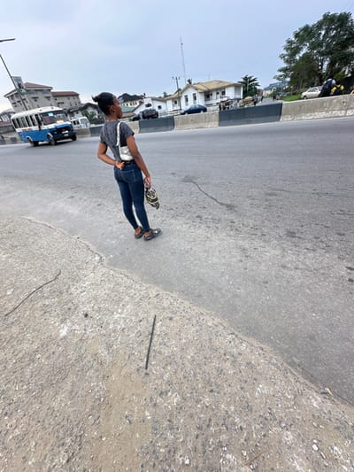 A young woman stands on a street, wearing jeans, a gray shirt, and dark sneakers. She carries a patterned bag. The street appears to be in a residential or commercial area with buildings and vehicles visible in the background. The scene is bright, with a clear sky. A thin, dark object lies on the sidewalk near the edge of the street. The overall impression is of an ordinary day on a busy street.