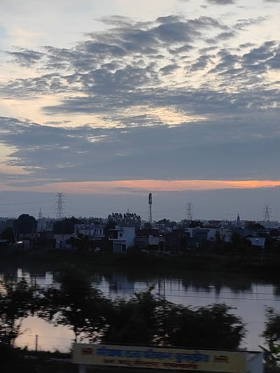 The image depicts a rural or semi-urban landscape at dawn or dusk, likely near Samana Bahu, India, given the context provided. A body of water, possibly a river or canal, occupies the foreground, reflecting the sky and the structures on the opposite bank. The water is dark and appears still, with some ripples and reflections of trees and buildings.
The middle ground features a line of buildings, appearing to be houses and smaller structures, interspersed with trees and vegetation. Several prominent electricity pylons with high-voltage power lines are visible in the distance, indicating the presence of infrastructure. One taller tower, possibly a communication mast, stands out amongst the buildings. The architecture suggests a modest settlement.
The sky above is dramatic, with a layer of textured, greyish clouds forming patterns. Towards the horizon, there is a soft band of orange and pink light, characteristic of sunrise or sunset. The overall lighting is subdued, creating a somewhat tranquil yet slightly somber atmosphere. No people are discernable in the image. The scene conveys a sense of a quiet, undeveloped area on the outskirts of a populated region.