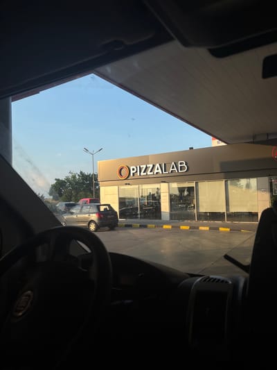 The image depicts a scene observed from inside a vehicle, likely parked at a service station in Tsaratsovo, Bulgaria. The viewpoint is from the driver's perspective, with a dark steering wheel displaying a Fiat logo and dashboard elements visible in the foreground.
In the midground, a modern, single-story commercial building features the prominent white signage "PIZZALAB" with an orange and grey circular logo to its left. The building has large glass windows, some of which are covered by interior blinds, revealing tables and chairs inside. A "NO SMOKING" sign is affixed to the right side of the building facade. The building is situated adjacent to an asphalt paved area, marked by a yellow and black striped curb. Distinct tire marks are visible on the asphalt.
Several vehicles are present in the paved area: a grey Skoda Fabia hatchback, displaying a Bulgarian license plate "A 9949 KK", is parked facing away from the camera. Partially obscured behind it is a dark-colored sedan. Further in the background, a red vehicle is visible near a line of green trees and shrubs.
The scene is under a clear, light blue sky, suggesting late afternoon or early evening light. A tall, dark street light pole stands to the left of the PIZZALAB building. A large, light-colored canopy structure, characteristic of a fuel station, extends over the building and portions of the paved area. Beyond the immediate commercial structures, a natural landscape with trees and a hazy horizon is visible.
