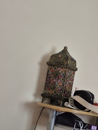 An ornate, antique-style lantern with multicolored glass inlays and a metallic filigree design sits on a wooden shelf. The lantern is positioned on the right side of the image, leaning slightly. Below the lantern, also on the shelf, are a black zippered pouch with a white strap bearing the word "GUESS" and a white tube. A black wire extends from the shelf downwards. The shelf itself is supported by a metal structure. The background is a plain, light-colored wall with a shadow cast by the lantern. The scene appears to be an indoor setting, likely a room within a dwelling. There are no people or animals visible in the image.