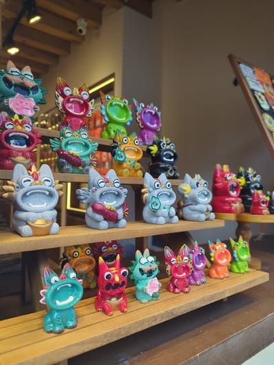 The image shows a close-up, eye-level shot of a display of colorful ceramic dragon figurines, likely from a shop in Dali City, China. The figurines are arranged on wooden shelves, with the foreground featuring a row of smaller dragons and the background showing larger ones. The dragons are depicted with open mouths, some holding golden ingots or red fish. The overall lighting is warm and inviting, highlighting the vibrant colors and intricate details of the figurines. The shelves are made of light-colored wood, and the background appears to be an indoor setting with some lighting fixtures visible on the ceiling. A framed board with what appears to be artwork or product information is visible in the far right background.