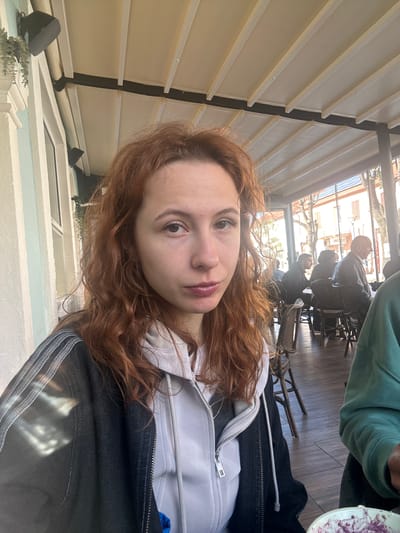 The image is a selfie taken at an outdoor cafe in what is possibly Nikšić, Montenegro. The foreground features a young woman with red curly hair wearing a light gray hooded sweatshirt and a black jacket. In the background, there are people seated at tables. One person is wearing a green jacket. The tables are on a wooden floor under a covered outdoor area with a white paneled roof structure. Outside the cafe, buildings are visible. There are speakers mounted on the outside wall of the cafe, and there is a small decorative shrub.