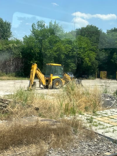 A yellow backhoe loader is parked on a concrete lot surrounded by dry grass and trees. In the background, a large, textured mural is visible on a wall, depicting abstract figures. Further back, there are more trees, a few parked cars, and yellow metal structures that resemble fences or cages. The sky is blue with scattered white clouds. This scene is located at the abandoned "Pioneers' Palace" (Palace of Young Pioneers) in Plovdiv, Bulgaria.