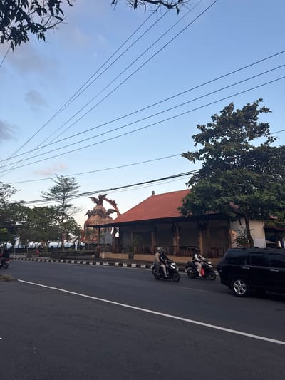 In Dawan, Indonesia, an overcast sky looms over a roadside scene. A dark-colored SUV is parked on the right side of a paved road, its passenger side wheel visible. Two motorcycles are traveling on the road in the same direction as the SUV. The first motorcycle carries two individuals wearing helmets. The second motorcycle is ridden by a single person, also wearing a helmet.
In the background, a building with a red tiled roof and an open-air veranda is situated behind a low wall. A large, imposing statue, appearing to be a mythological creature with wings and sharp features, stands behind the building. Trees are scattered throughout the scene, with some foliage in the foreground and more densely packed trees lining the area behind the wall. Electrical wires are strung across the sky, adding a linear element to the composition. The road itself is marked with a white line, dividing the lanes of traffic.