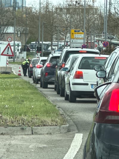 The image shows a line of cars stopped in traffic in Kehl, Germany. A person wearing a high-visibility vest is standing near traffic cones on the right side of the road. Road signs indicate the direction to "Zentrum" (center) and other locations. A speed limit sign with a "10" is visible. The license plate of the second car in line begins with "G".