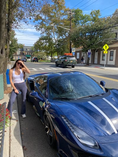 An eye-level outdoor scene in Ridgewood, United States, depicts a young female subject standing on a concrete sidewalk next to a parked dark blue Ferrari convertible, which features two white racing stripes on its hood. The female, possessing long dark hair, wears sunglasses, a white halter top, dark flared pants, and white sneakers. She holds a smartphone in her left hand, with her right hand raised near her forehead, and is looking towards the camera. The driver's side door of the Ferrari is open. To the left, a partial view of a metal bench, a blue trash receptacle, and red flowers in a planter is visible.

The background shows a two-lane street with a double yellow line. A dark green Toyota 4Runner SUV, fitted with a roof rack carrying an orange kayak or similar item, travels away from the camera on the right side of the road. Other vehicles are present further in the distance. The street is lined with mature trees displaying green and some autumnal foliage. Buildings with brick and stone facades, windows, and awnings flank the road, featuring a yellow diamond-shaped pedestrian crossing sign and a "NO PARKING" sign on the right. Overhead utility lines and wooden poles are visible against a clear blue sky.