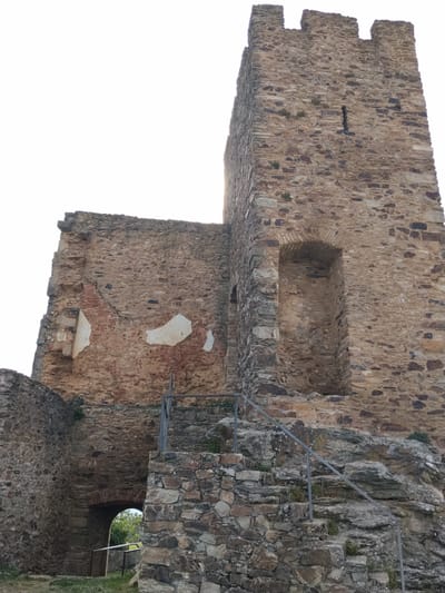 This is a low-angle, eye-level, outdoor shot of a ruined castle tower in Mogadouro, Portugal. The tower, constructed of irregularly shaped, brown and tan stones, rises high in the frame. The top of the tower has a crenellated parapet. A vertical slit window is visible near the top of the tower.
Adjacent to the tower, a lower section of the castle ruins, also constructed of similar stones, has areas of exposed brick and whitewashed sections. There is a recessed arched opening with a stone border in the tower. Steps lead up to this opening, with a gray metal railing running alongside them.
At the base of the ruins is a low, arched stone entrance. Green vegetation is visible through this entrance. The ground in the foreground is covered with grass and a few small plants. The sky is overcast and a bright white.