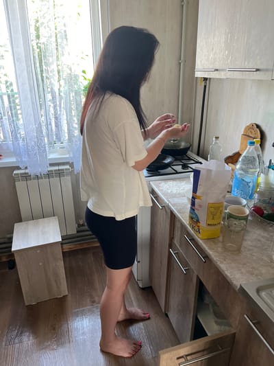 A woman with dark hair, wearing a white t-shirt and black shorts, stands in a kitchen, preparing food. She is holding small, round objects in her hands. The kitchen has light wood cabinets, a countertop with various items including a bag of flour and bottles of water, and a gas stove. A window with sheer curtains is visible in the background, allowing natural light to illuminate the scene. A white radiator is located beneath the window, and a small wooden stool is placed next to it. The floor is made of dark wood planks. The overall impression is of a domestic setting, possibly a home kitchen in Navahrudak, Belarus, given the context.