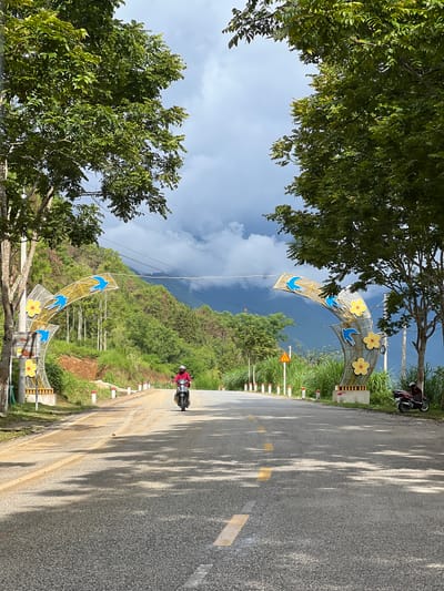 A wide-angle, eye-level shot captures a paved road in Quản Bạ Commune, Vietnam, framed by lush green trees on either side. The road slopes gently upwards and narrows into the distance, with yellow dashed lines marking the lanes. On the left and right sides of the road, decorative arches with yellow flowers and blue bird motifs are installed. A person on a scooter, wearing a red shirt and helmet, is traveling away from the camera on the right side of the road. In the background, a verdant, mountainous landscape is visible under a cloudy sky. A parked scooter is situated on the right side of the road near the decorative arch.