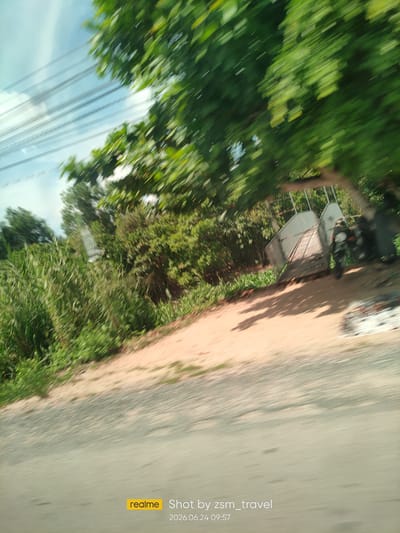 This is a motion-blurred image taken on a sunny day in Ben Cau, Vietnam. The scene depicts a dirt road bordered by lush green foliage. In the background, a metal gate is visible, partially obscuring trees and vegetation. A motorcycle is parked near the gate. The sky is a light blue with some white clouds. The timestamp on the image indicates it was taken on June 24, 2026, at 09:57. The presence of overhead power lines suggests a rural or semi-urban area.