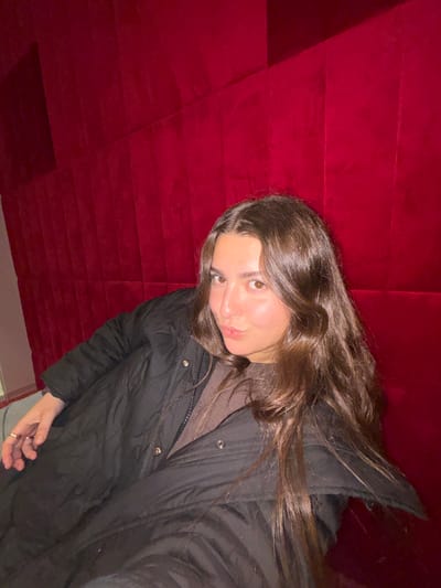 A young woman with long brown hair and brown eyes wearing a brown shirt and a black jacket is angled towards the camera. She is laying back, perhaps on a red velvety wall, with her hair partly covering her face. Her right hand is visible near the bottom corner, displaying a ring. The background is a textured, possibly padded, red surface.