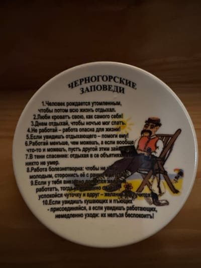 The image displays a white, round ceramic plate with text and an illustration. The text is in Russian and appears to be a list of humorous rules or advice. The title at the top reads "ЧЕРНОГОРСКИЕ ЗАПОВЕДИ" which translates to "MONTENEGRIN COMMANDMENTS". Below the title, there are numbered points, some of which are visible and partially legible, starting with "1. Человек рождается утомленным..." (1. A person is born tired...). An illustration features a cartoonish man with a mustache and hat, wearing a vest and sitting in a lounge chair on a yellow background, possibly representing a sunny day with a beach. The plate is angled and appears to be resting on a wooden surface. The lighting suggests indoor photography. There is no discernible connection to Sergeevka, Russia.