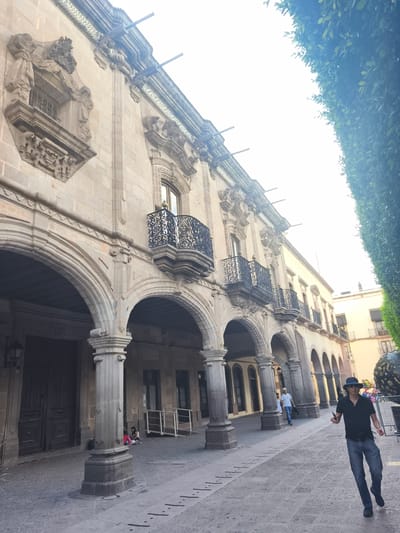The image depicts a street scene in Querétaro, Mexico, showcasing a building with colonial architecture, including arches, columns, and ornate balconies.
A man in a hat, black shirt, and jeans walks towards the camera on the paved street. Further down the street, two other people are walking away. A couple of children are sitting on the ground near a large column. An ornate, decorative sphere is visible at the edge of the frame, fenced off. The architecture indicates a historical building or area.