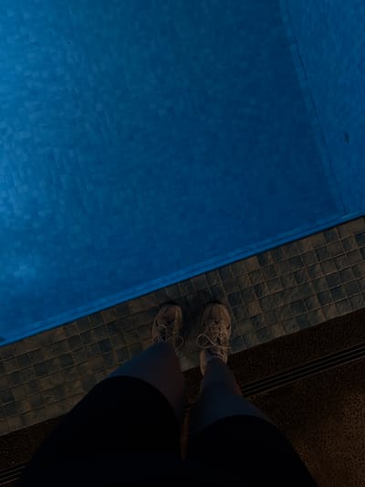 The image shows a portion of a swimming pool tiled in blue, with a person standing on the tiled deck next to it. The person is wearing athletic shoes and dark shorts. The deck has a dark tile border. The location appears to be indoors due to the controlled lighting. The picture was presumably taken in Jinhua, China as requested.