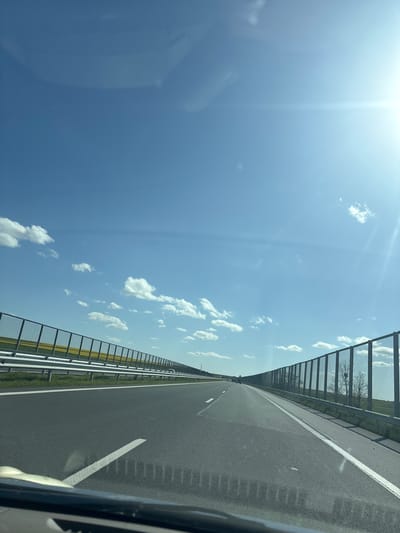 The image captures a bright, sunny daytime scene from the perspective of a vehicle moving on a multi-lane highway, likely in Chokoba, Bulgaria. The photo is oriented ninety degrees clockwise, with the road appearing on the right and the sky on the left.

On the right side of the rotated image, a well-maintained asphalt highway stretches into the distance, marked with clear white dashed and solid lines. A white solid line separates the main lanes from a wide shoulder. Parallel to the road, a metal guardrail or sound barrier extends along the side. Beyond this barrier, green vegetation is visible, leading to extensive yellow fields, characteristic of blooming rapeseed or similar crops.

The left side of the image is dominated by a clear, vibrant blue sky adorned with scattered, fluffy white cumulus clouds. The bright sun is prominently featured in the upper left corner, creating a strong lens flare and streaks of light that radiate across the sky, indicating intense daylight. The overall weather appears fair and bright.

The very edges of a vehicle's interior, specifically a part of the dashboard or windshield frame, are visible in the bottom right corner and subtly in the top left, confirming the photograph was taken from inside a car in motion. There are no people visible, and no discernible text appears in the image.