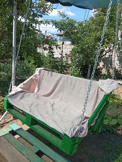 A homemade swing made from green-painted wooden pallets is covered with a light-colored blanket. The swing is suspended by metal chains from a support structure, partially obscured by a green mesh shade. The scene is set in a garden with lush green foliage, including trees with visible fruits and some pink flowers. In the background, a brick wall and parts of buildings suggest a residential area. The ground is covered with soil and scattered vegetation. The sky is blue with scattered clouds. The location is identified as Tomai, Moldova.
