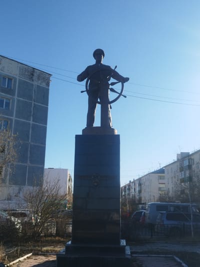 The image shows a bronze statue of a sailor holding a ship's wheel, erected on a dark stone pedestal in Zhatay, Russia. The statue is centered in the frame, with the clear blue sky as its backdrop. Buildings of typical Soviet-era architecture surround the statue on both sides. A few bare trees and bushes stand in the foreground, partially obscuring the view of parked cars and a paved sidewalk surrounding the monument. Power lines cross above the monument.