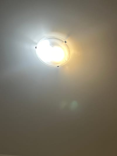 The image is a close-up, low-angle shot of a ceiling light fixture in Richmond Hill, Canada. The fixture is circular with a translucent cover, and a bright light bulb illuminates the ceiling. The light is a mixture of white and yellow, suggesting a warm tone. The ceiling is painted a neutral gray color, and the texture of the paint is visible. There are two small metallic clips holding the fixture in place, visible on the left and right sides of the light. A faint greenish lens flare is visible in the lower right quadrant of the image. The overall scene is unremarkable and appears to be a typical residential or commercial indoor setting.