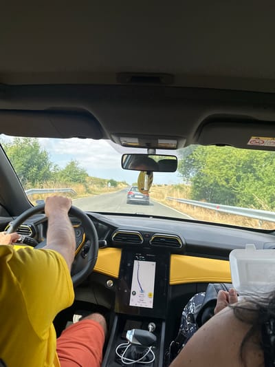 The image shows the interior of a car driving on a road in a rural area with trees and dry grass on the sides. The driver, wearing a yellow t-shirt and orange shorts, has their hands on the steering wheel. The car's dashboard features a large touchscreen display showing navigation, and yellow accents. In the passenger seat, a person wearing patterned leggings and a black jacket is visible, holding a white container. The road ahead shows another car and a guardrail. The image is taken from the perspective of someone in the passenger seat, looking forward. The scene suggests a journey on a sunny day.