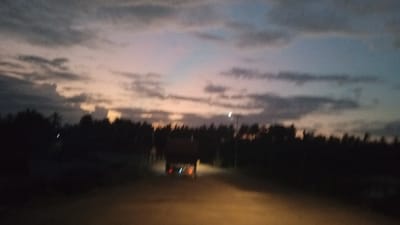 The image depicts a dimly lit, blurred scene of a road at dusk, likely in Awegeutahpaya, Indonesia. The sky is filled with muted colors of gray, pink, and blue, suggesting a sunset or twilight. A silhouette of a truck is visible on the road, its taillights illuminated. Dark trees line the sides of the road, obscuring any further details of the surrounding environment. The overall impression is one of a quiet, possibly rural, setting.
