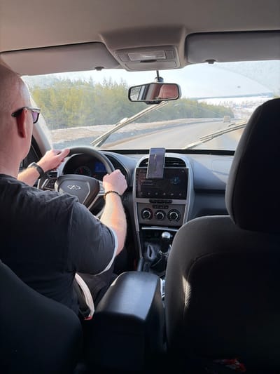 The scene is captured from the interior of a vehicle, looking forward from a rear passenger seat on a sunny day in Zabegalovo, Russia. A bald, male driver, wearing sunglasses and a dark short-sleeved shirt with white trim, operates the car, his hands positioned on the steering wheel. A black digital watch is on his left wrist and a dark beaded bracelet on his right. The car's dashboard features a central infotainment screen, climate controls, and a smartphone mounted vertically above the central vents, displaying a navigation application. The instrument cluster shows the vehicle speed at approximately 60-70 units (likely km/h). A manual transmission gear stick is visible in the center console. The windshield is noticeably dirty with streaks, and the wipers are at rest at the bottom. Through the windshield, a two-lane paved road is visible, curving to the right. The road shoulders are partially covered in snow. A dense evergreen forest lines the left side of the road. In the distance on the right, a snowy, open landscape with scattered buildings or a settlement is discernible under a clear sky. A reflection, likely of the driver's head, is visible in the rearview mirror. A beige seatbelt buckle is seen unbuckled between the driver's seat and the center console armrest.